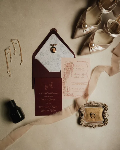Invitation suite wedding invitation flat lay with burgundy envelope, wax seal, pearls, perfume, ring, and satin bow heels on neutral fabric backdrop