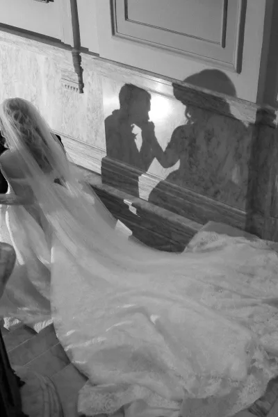 Ceremony moment with bride and groom holding hands in church pews, bridal veil trailing as sunlight casts dramatic shadows on marble wall
