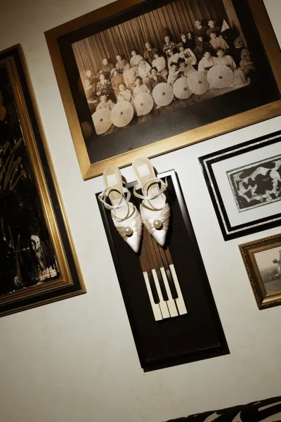 Wedding shoes with pearl embellished white satin heels and ankle straps displayed on a gallery wall with framed vintage photos