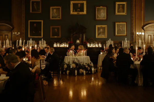 Wedding reception decor with tall candelabras and taper candles, red floral centerpieces, and white linens in a historic portrait-lined hall