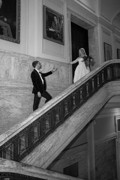 Wedding first look as groom in tuxedo waits on a marble staircase, reaching for the bride in veil and strapless gown entering the doorway