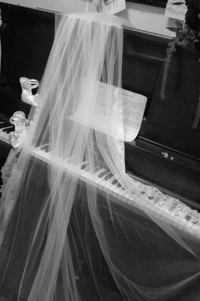 Bridal veil draped over an upright piano with lace trim accent, beside bridal shoes, wedding rings, and sheet music on hardwood floor