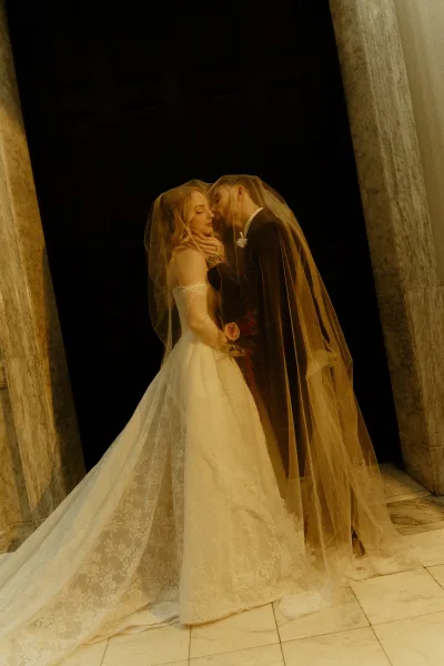 Wedding kiss portrait with veil draped over couple as groom kisses bride’s forehead beside stone columns and a dark doorway