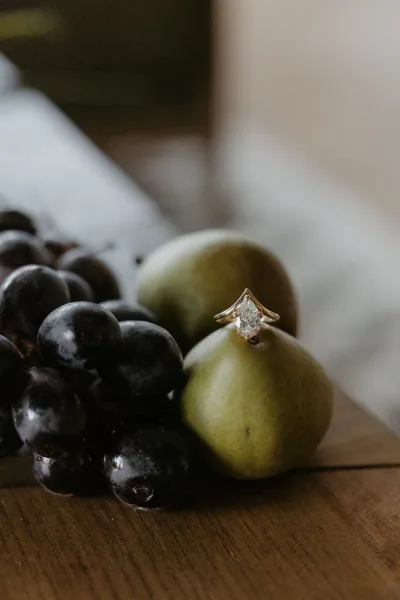 Engagement ring with a pear shaped diamond ring on grapes and a pear atop a wooden tabletop against a soft neutral backdrop
