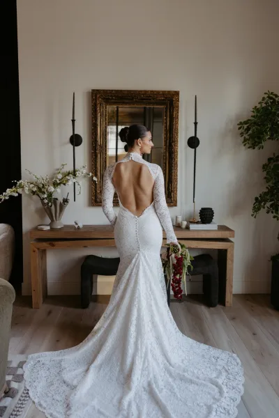 Bridal portrait of a bride in a backless wedding dress, looking over her shoulder with a cascading bouquet beside a framed mirror indoors