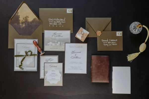 Invitation suite wedding stationery flat lay with kraft envelopes, wax seal, green velvet ribbon, rings and vow books on black fabric backdrop