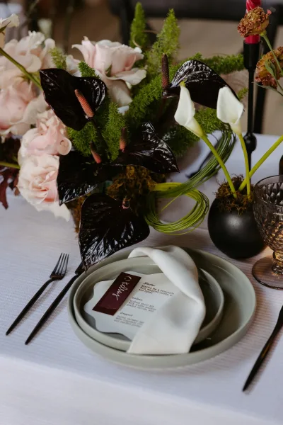 Reception tablescape with wedding place setting, anthurium and calla lily centerpiece, black taper candle, and smoked goblet with blurred seating behind