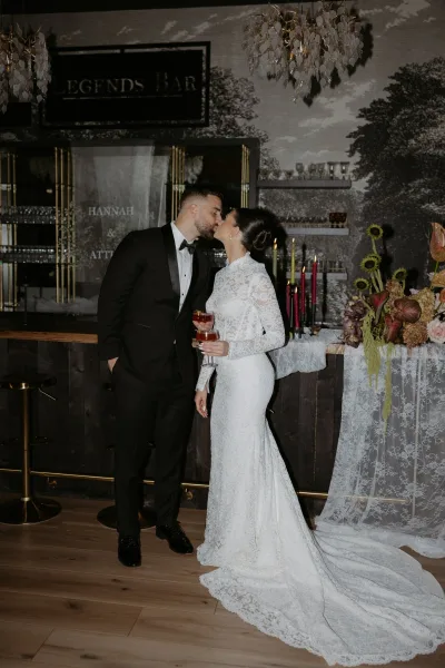 Wedding kiss portrait of bride and groom kissing at an indoor bar, bride in long-sleeve lace dress holding a cocktail under a chandelier with red taper candles