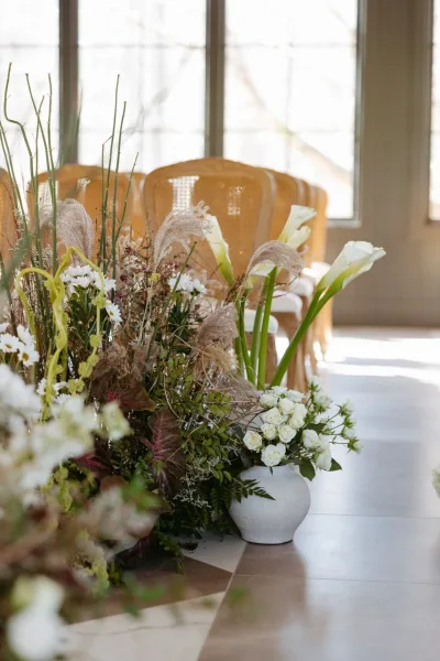 Ceremony aisle florals with a wedding aisle flower arrangement of white calla lilies, roses, daisies, and pampas grass by rattan chairs in window-lit room