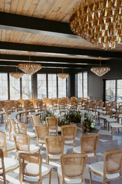 Ceremony setup with circular cane-back chairs and white cushions around a floral meadow, under chandeliers in a windowed indoor venue