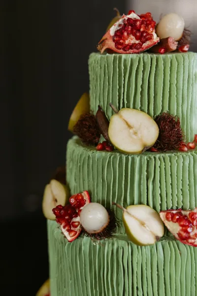 Wedding cake with green wedding cake buttercream texture, topped with pomegranate, pear slices, rambutan, and lychee against a dark backdrop