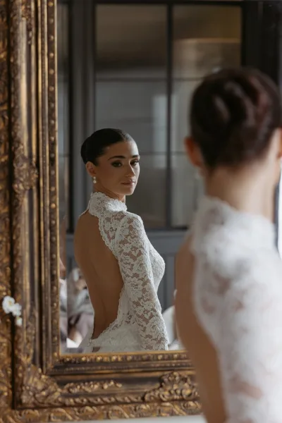 Bridal portrait of a bride in mirror wearing a backless lace, high-neck long-sleeve gown and pearl drop earrings beside an ornate gold frame.