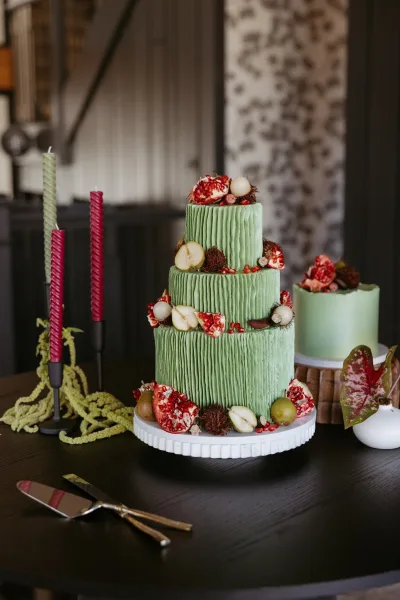 Wedding cake with green frosted tiers topped with pomegranate and pear slices, surrounded by candles and greenery on a dark wood table
