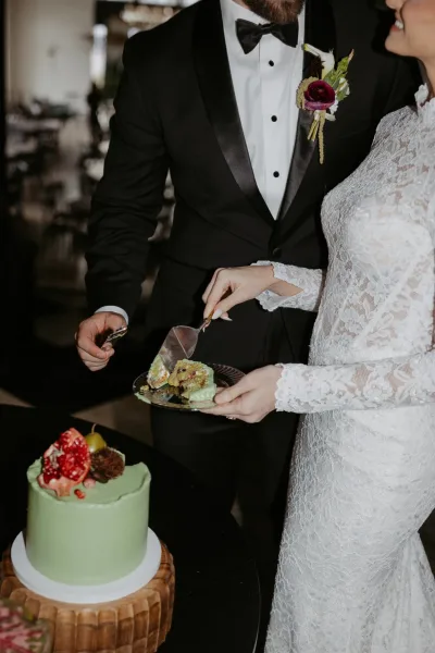 Wedding cake cutting as bride in lace long sleeves holds a dessert plate while groom in black tuxedo uses server at reception table