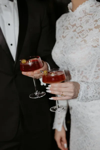 Wedding cocktail toast as bride and groom clink red coupe drinks with olive picks, lace sleeve and rings visible against a dark interior backdrop