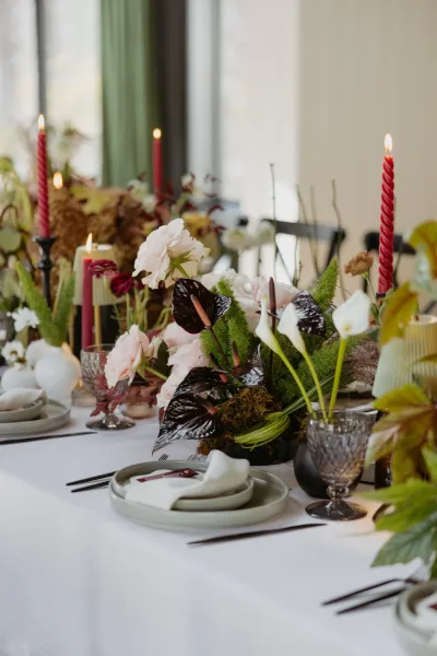 Reception tablescape with wedding table centerpiece of anthuriums, calla lilies, blush roses, and red taper candles by curtained windows