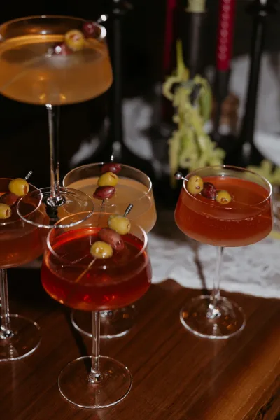 Wedding cocktails in coupe glasses with olive picks on a wood table, liquor bottles behind in a dim bar setting