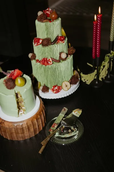 Wedding cake with green wedding cake tiers in textured buttercream, topped with figs, pomegranate, pear and grapes beside red taper candles in a dark room