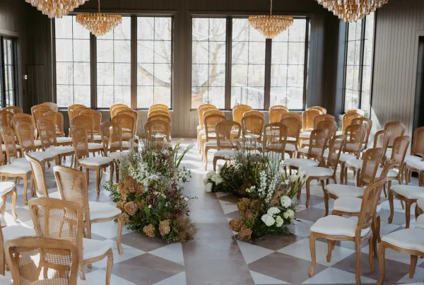 Ceremony setup for an indoor wedding ceremony with cane back chairs, white cushions, and aisle florals in a windowed room with chandeliers