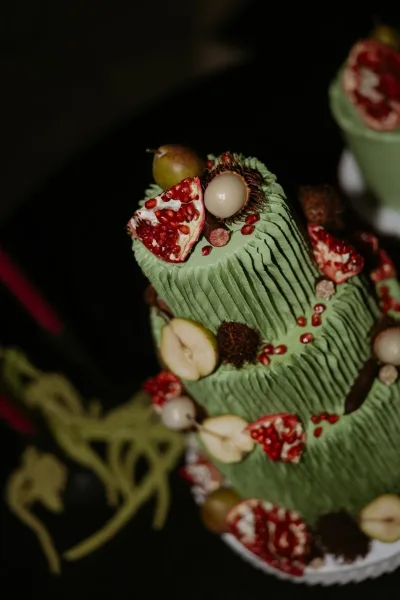 Wedding cake with green wedding cake textured buttercream, topped with pomegranate, pears, and rambutan on a cake table against dark backdrop