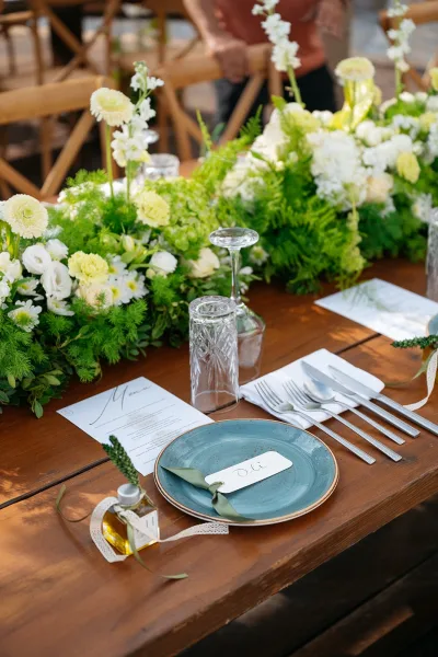 Wedding place setting on a rustic wood reception table in an outdoor setting, featuring a petite olive oil favor and nearby wooden chairs.