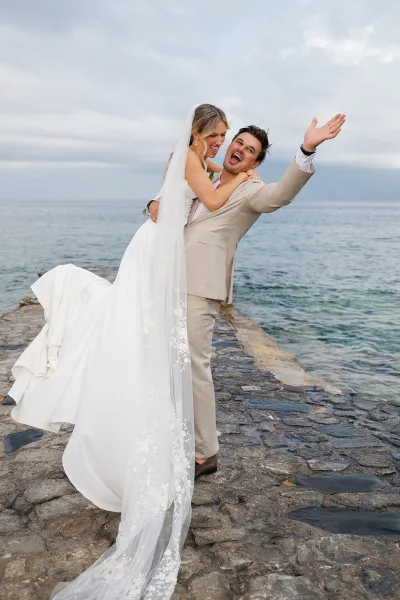 Couple portrait with the groom lifting the bride on a stone pier by the ocean, her long veil catching the breeze beneath a cloudy sky