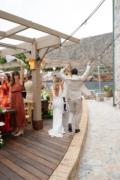 Wedding reception entrance as the bride and groom arrive, guests wave napkins on a waterfront deck against rocky cliffs under an overcast sky.