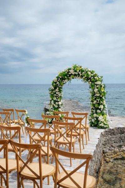 Ceremony setup featuring a floral wedding arch and crossback chairs on a stone pier by the ocean under a soft, cloudy sky.