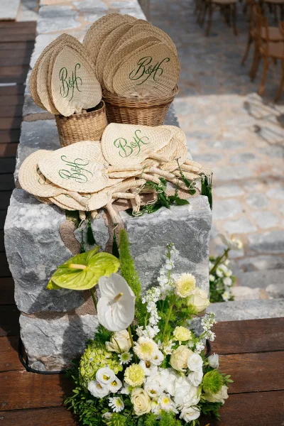 Wedding fans arranged as hand fan wedding favors with monogram embroidery, neatly displayed on a wood deck against a rustic stone wall.