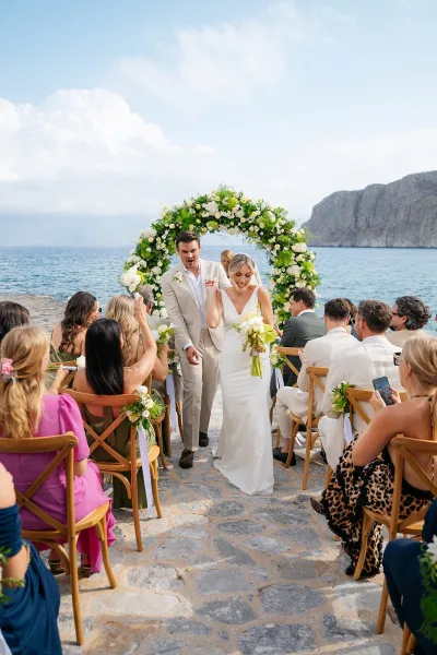 Ceremony recessional as the couple makes a just married exit beneath a circular green-and-white floral arch on a stone oceanfront walkway.