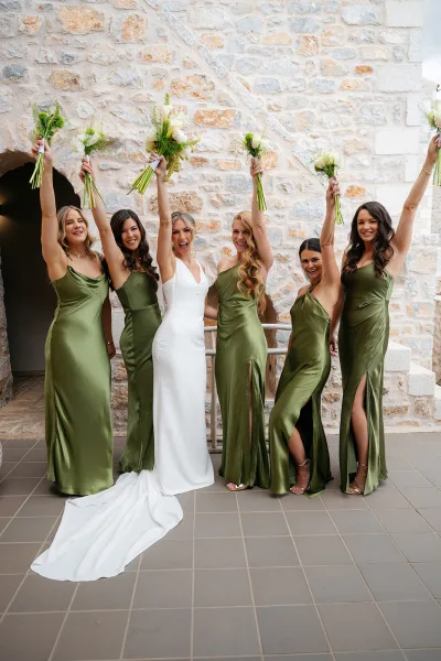 Bride and bridesmaids in olive green bridesmaid dresses hold greenery bouquets by an archway, the bride in a sheath gown.