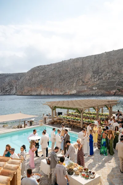 Wedding cocktail hour under a pergola for a poolside cocktail hour, overlooking the ocean and rocky cliffs on a stone patio.