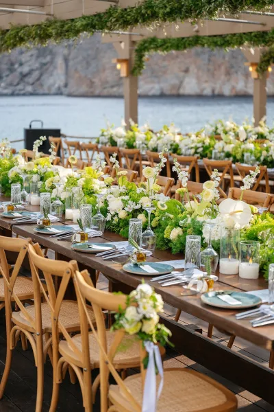 Reception tablescape with long banquet table decor, candles in glass hurricanes, set on a wooden deck by the ocean beneath a pergola.