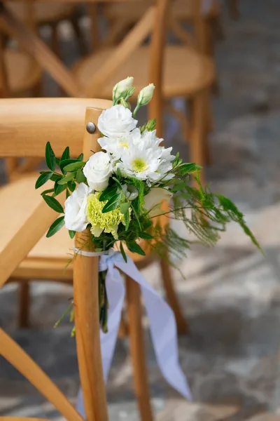 Aisle chair flowers tied with a white ribbon hang from a wooden chair as elegant ceremony aisle decor on a simple stone floor.