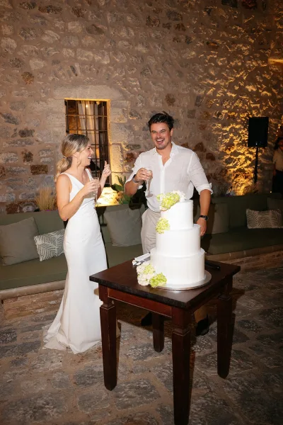 Wedding cake cutting as the bride and groom toast, slicing a three-tier white buttercream cake, champagne flutes by a rustic stone wall.