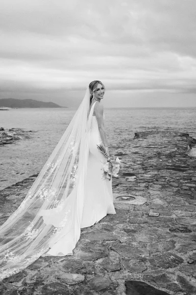 Black and white bridal portrait of a bride with long veil on a stone pier by the ocean beneath a cloudy sky, distant hills faintly visible.