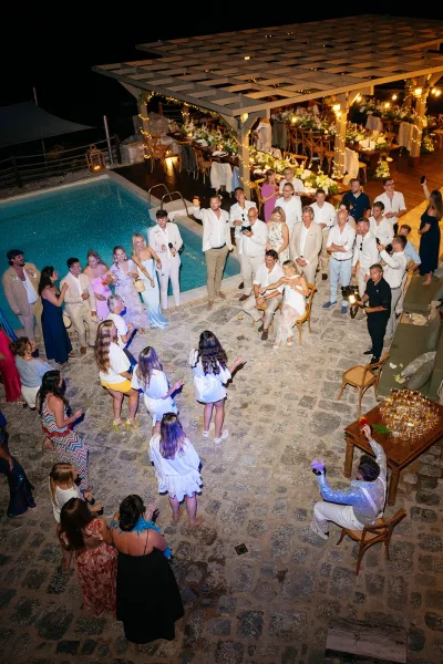 Reception dance party fills the wedding dance floor by the pool under string lights, with a pergola on the stone patio against the night sky.