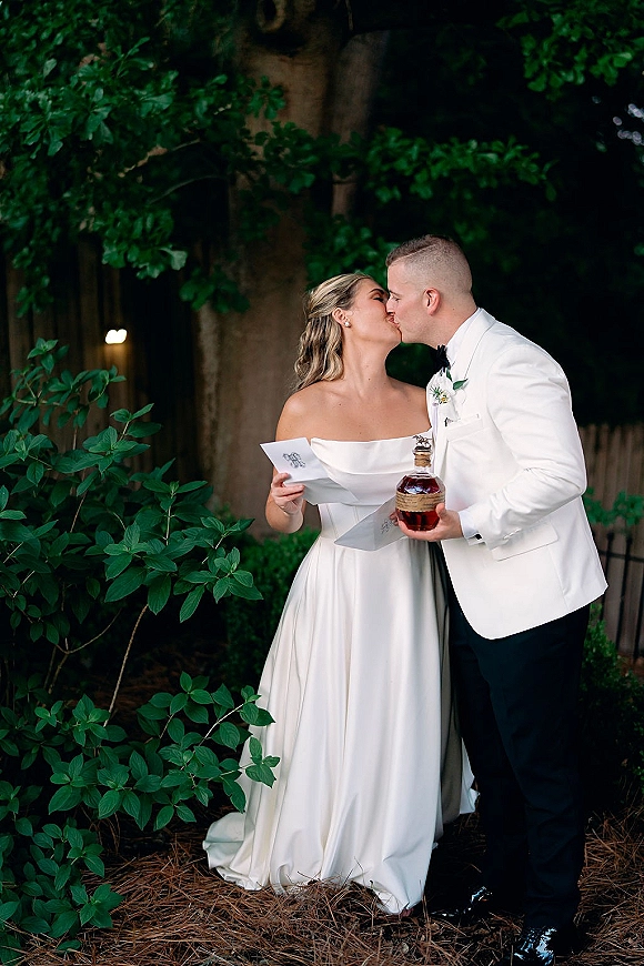 Wedding kiss portrait of bride and groom kissing, her off-shoulder gown and his white tuxedo, holding vow booklets in a garden setting