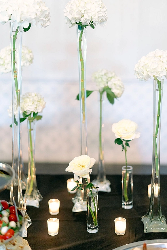 Wedding centerpiece with white hydrangeas and roses in tall clear glass vases, floating candles and tea lights on a dark tabletop