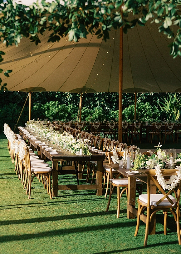 Reception tablescape at an outdoor tented reception with long wooden tables, white floral and greenery garlands, candles, and string lights on a lawn