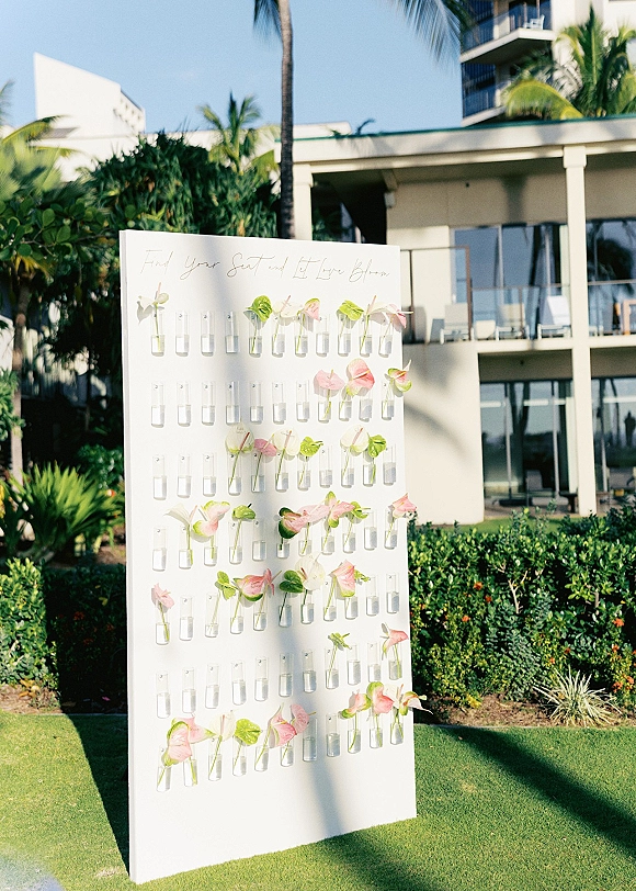 Wedding seating chart escort card display on a white board with acrylic name cards, bud vases, anthurium, and greenery on a tropical lawn backdrop