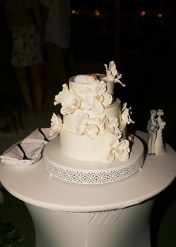 Wedding cake with white buttercream in three tiers, topped with figurine and sugar flowers on lace stand amid string lights in dark reception