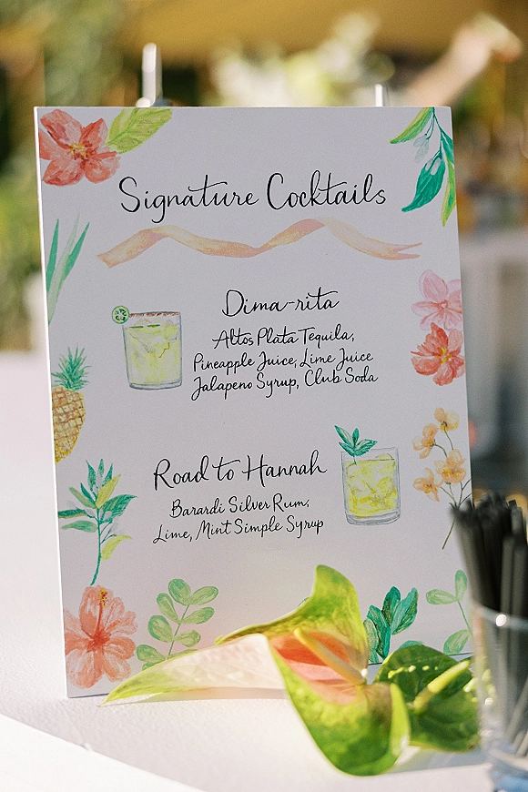 Signature cocktail sign wedding bar menu sign with calligraphy lettering, illustrated drinks and pineapple accents on a stand amid blurred greenery