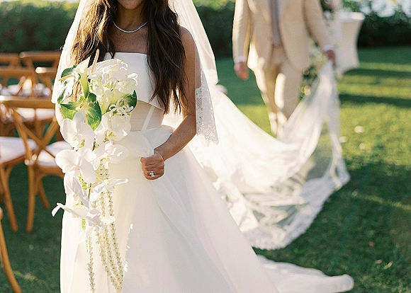 Bride portrait holding a white orchid bouquet with trailing garland, in a strapless dress and lace veil on a sunlit lawn aisle