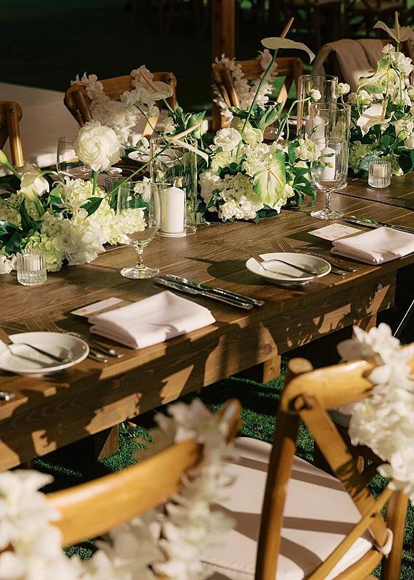 Reception tablescape on a farmhouse wedding table with white florals and greenery, cylinder vase candles, and place settings on a grass lawn