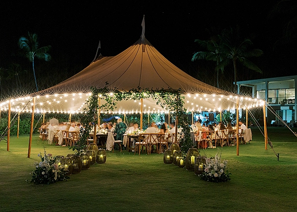 Wedding reception tent with sailcloth canopy, warm string lights, and long banquet tables with candles on a palm-lined lawn at night