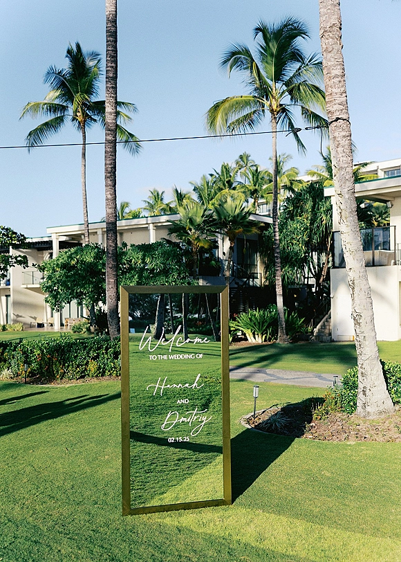 Wedding welcome sign on clear acrylic in a gold frame along a resort garden walkway with palm trees, lawn grass, and blue sky