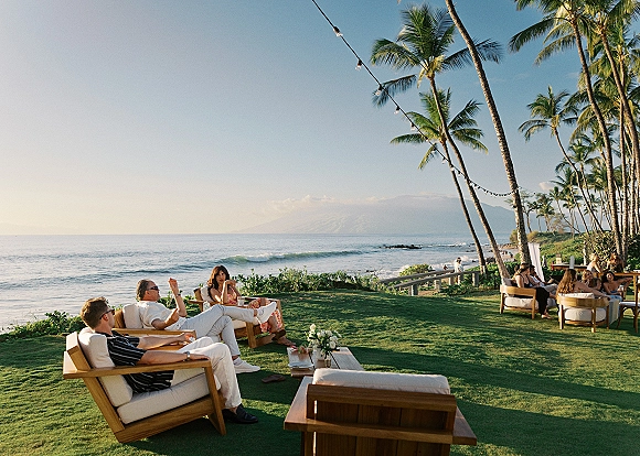 Wedding cocktail hour lounge seating with white outdoor sofas and wooden armchairs on a grassy lawn beside the ocean under string lights