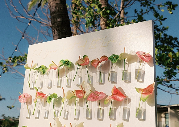 Seating chart display with calligraphy wedding seating chart on a white board, accented by anthurium flowers in clear bud vases under string lights