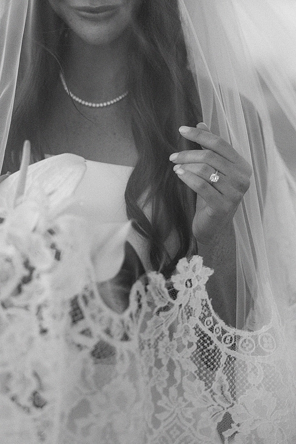 Bridal portrait of a bride holding veil with lace edging, pearl necklace, and bouquet against a neutral backdrop, showing her ring